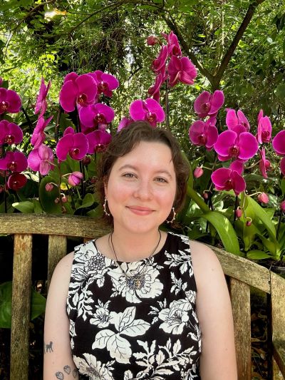 A woman is smiling as she sits outdoors in front of a bush adorned with vibrant purple flowers.